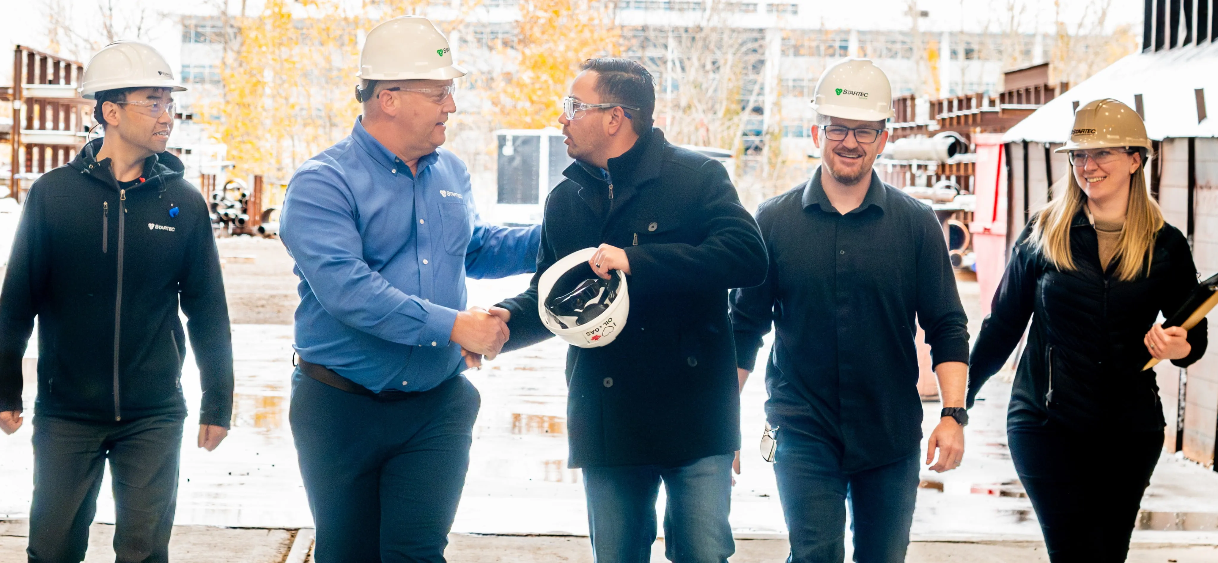 A picture of people walking together in a line in an industrial plant. There are two men shaking hands as they walk