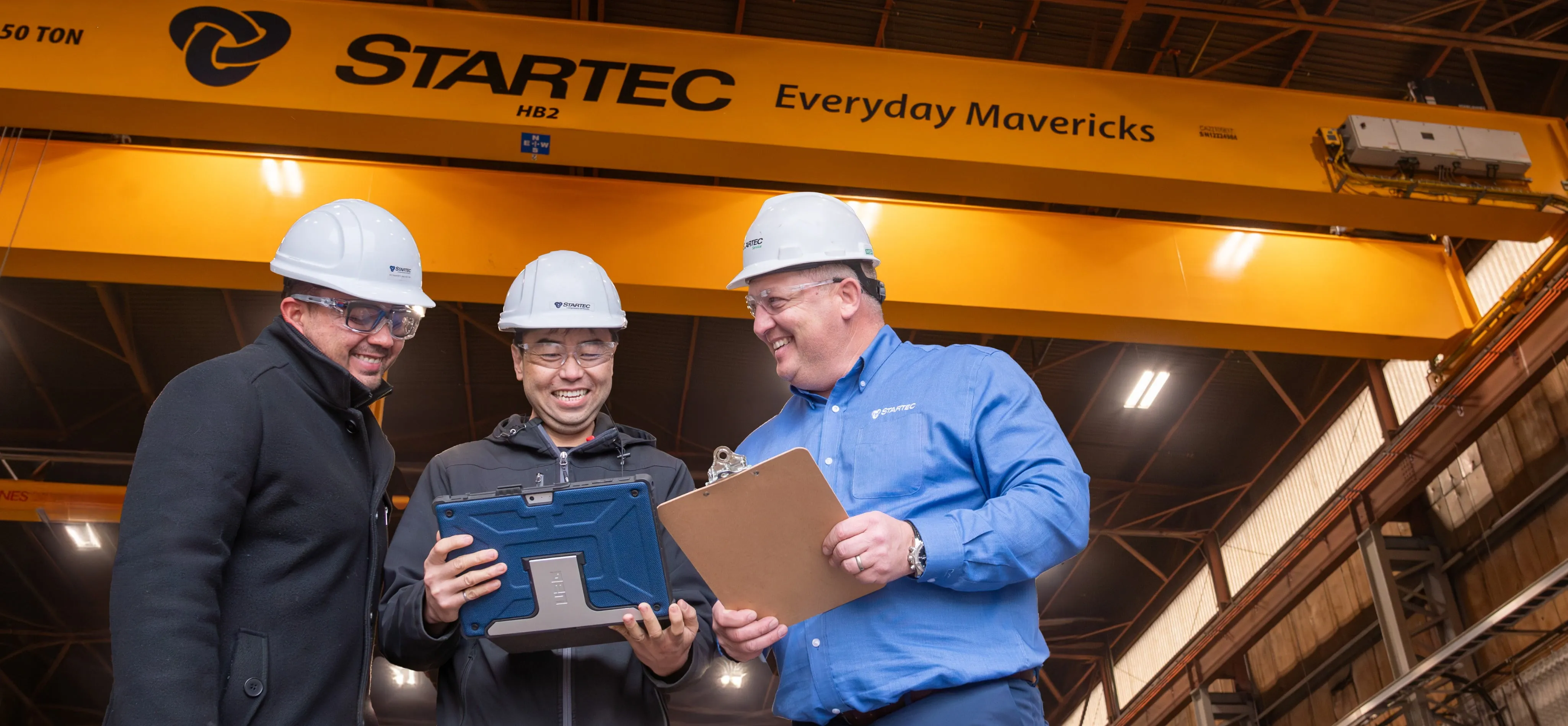 A picture of three men in hard hats having a discussion looking at a document inside a industrial plant