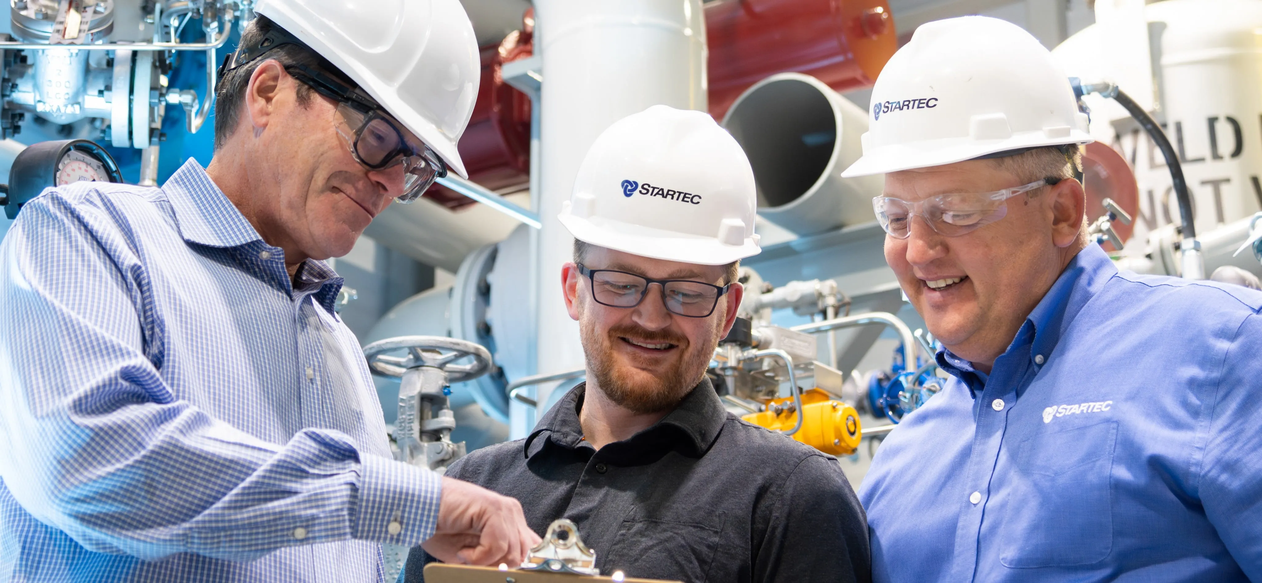 A picture of three men in hard hats having a discussion looking at a document inside a industrial plant